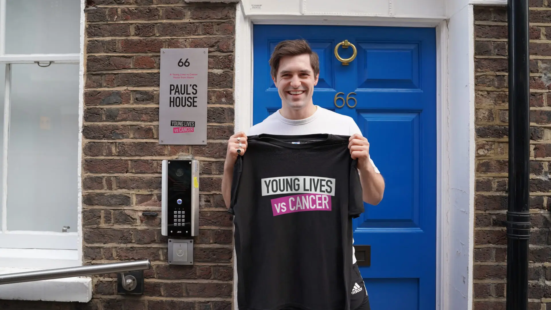 Phil Dunster is stood outside Paul's House, Young Lives vs Cancer's Home from Home. He is smiling and holding up a Young Lives vs Cancer t-shirt.